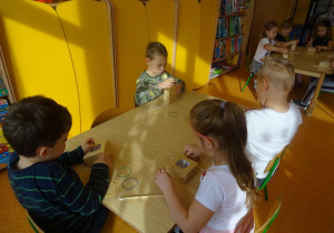 Czwórka dzieci siedzi przy stoliku, w rękach trzymają gumki recepturki, które zakładają na kartonowe małe pudełko.
